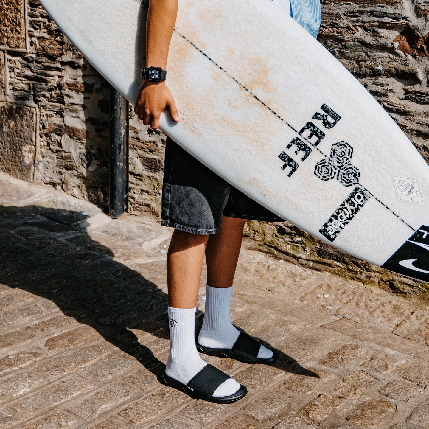 Person carrying a surfboard and wearing black open toe slides with toes facing right on a stone walkway