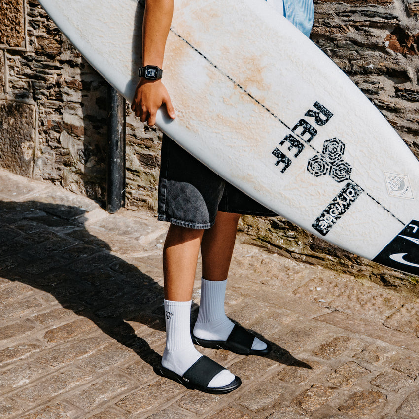 Person carrying a surfboard and wearing black open toe slides with toes facing right on a stone walkway