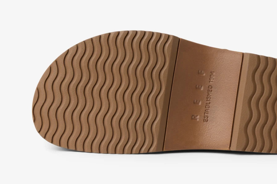 Close-up of tan sandal sole facing upward, showing wave-patterned rubber outsole and embossed REEF logo.